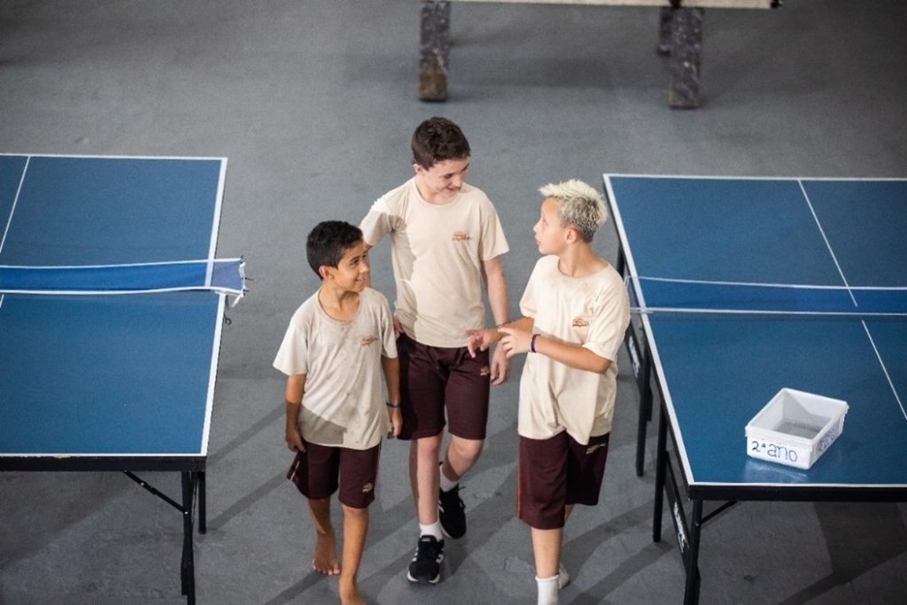 Três garotos uniformizados na escola andando entre duas mesas de ping-pong, para representar como decisões estratégicas dos gestores podem fazer os alunos mais felizes