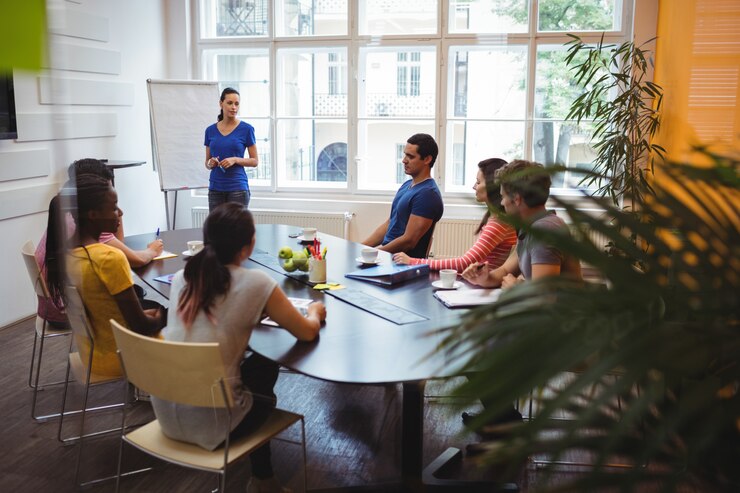 Equipe de colaboradores de uma empresa em uma mesa de reunião, com um dos integrantes de pé em frente a um quadro