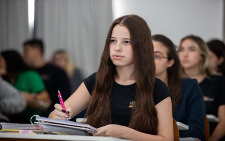 Menina com caderno e lapiseira sentada em sala de aula olhando para frente, com alunos no fundo, para exemplificar a importância da gestão escolar para o aprendizado
