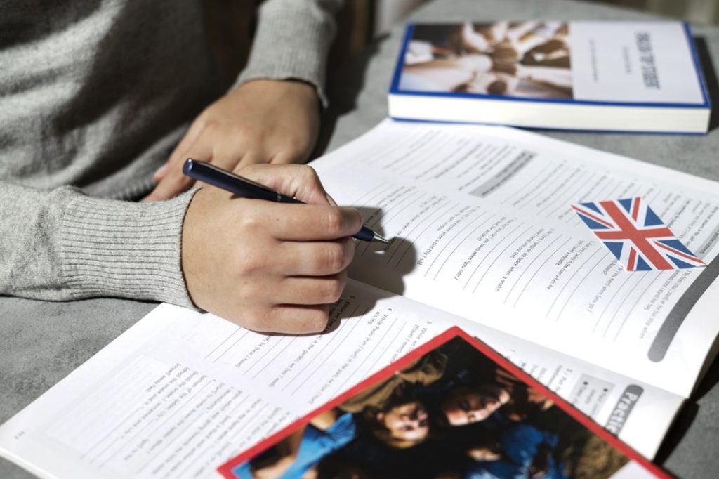 Mão com uma caneta escrevendo em um livro de atividades de inglês, para ilustrar a importância dos testes de proficiência